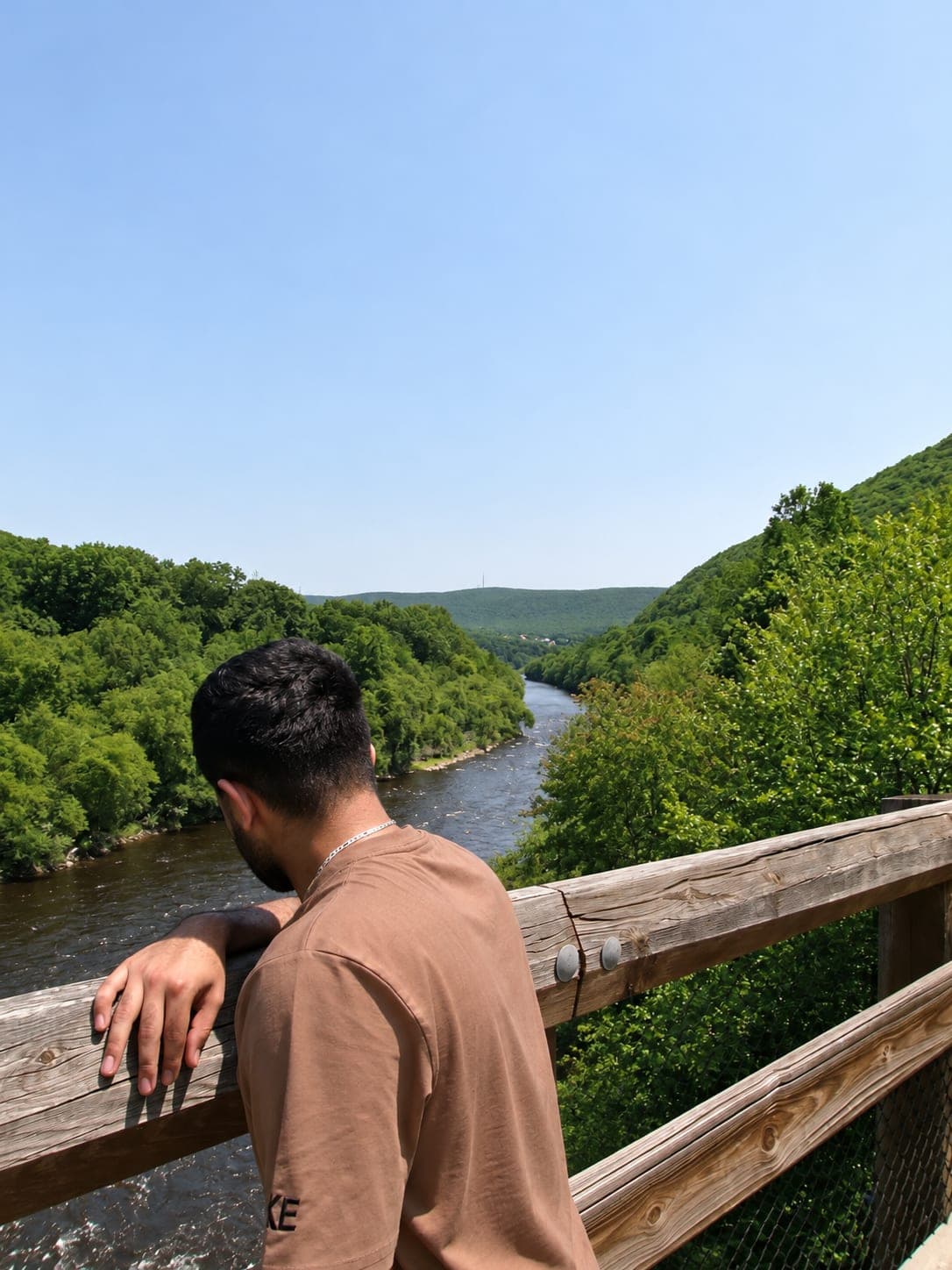 Sahil leaning on a wooden bridge railing above a river winding between green hills